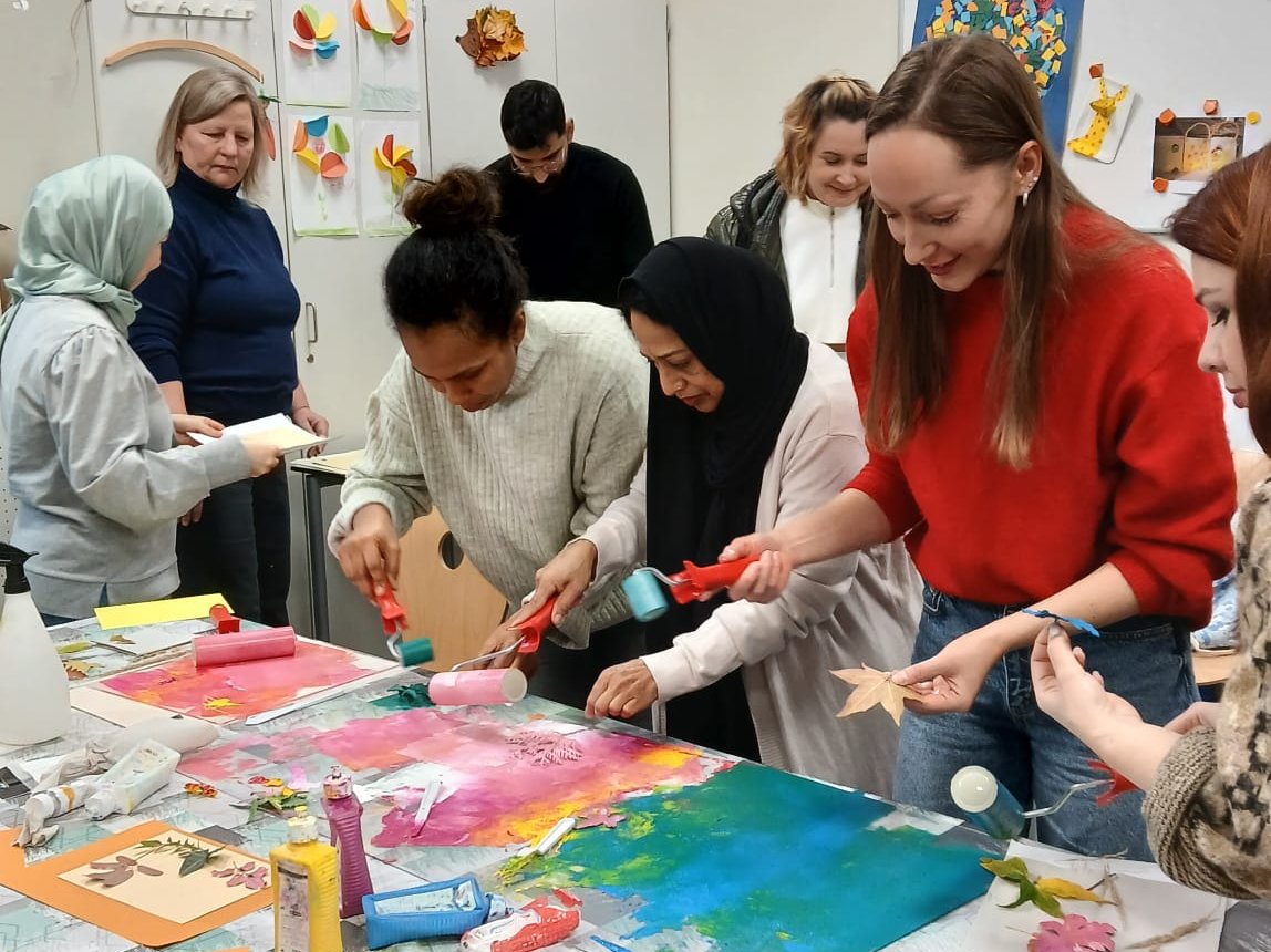 Eine Gruppe arbeitet in einem künstlerischen Prozess, es werden Pflanzenteile wie Blätter und Blüten in bunten Farben gedruckt. Im Vordergrund sieht man Blätter von verschiedenen Baumarten. Im Zentrum arbeiten mehrere Frauen sehr konzentriert an ihren Werken und im Hintergrund wird die Druckmaschine bedient.