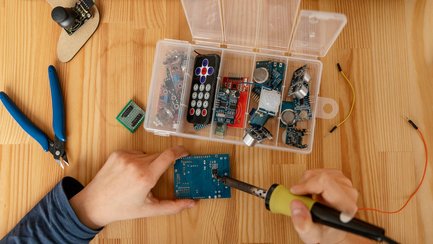 Quartierswerkstatt Elektro Person lötet Steckverbindungen auf einer Platine. Draufsicht auf die Hände und auf eine Ersatzteilkiste mit Dioden.