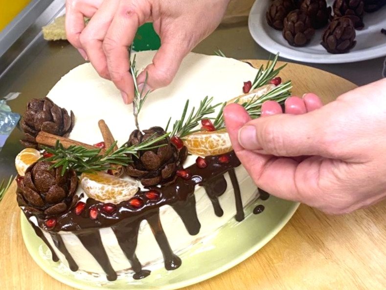 Eine weihnachtliche Torte wird verziert, man sieht Hände bei der Arbeit. Die Torte ist weiß und zum Teil mit einer Schokoladenglasur überzogen. Aus Corflakes wurde ein essbarer Tannzapfen hergestellt. Rosmarinzweige wirken wie zarte Tannenzweige.